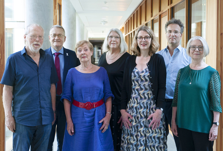 Auf dem Foto sehen wir die Mitglieder der Unabhängige Kommission zur Aufarbeitung sexuellen Kindesmissbrauchs nebeneinander in einem lichtdurchfluteten Büroflur stehend. Alle sieben schauen in die Kamera.