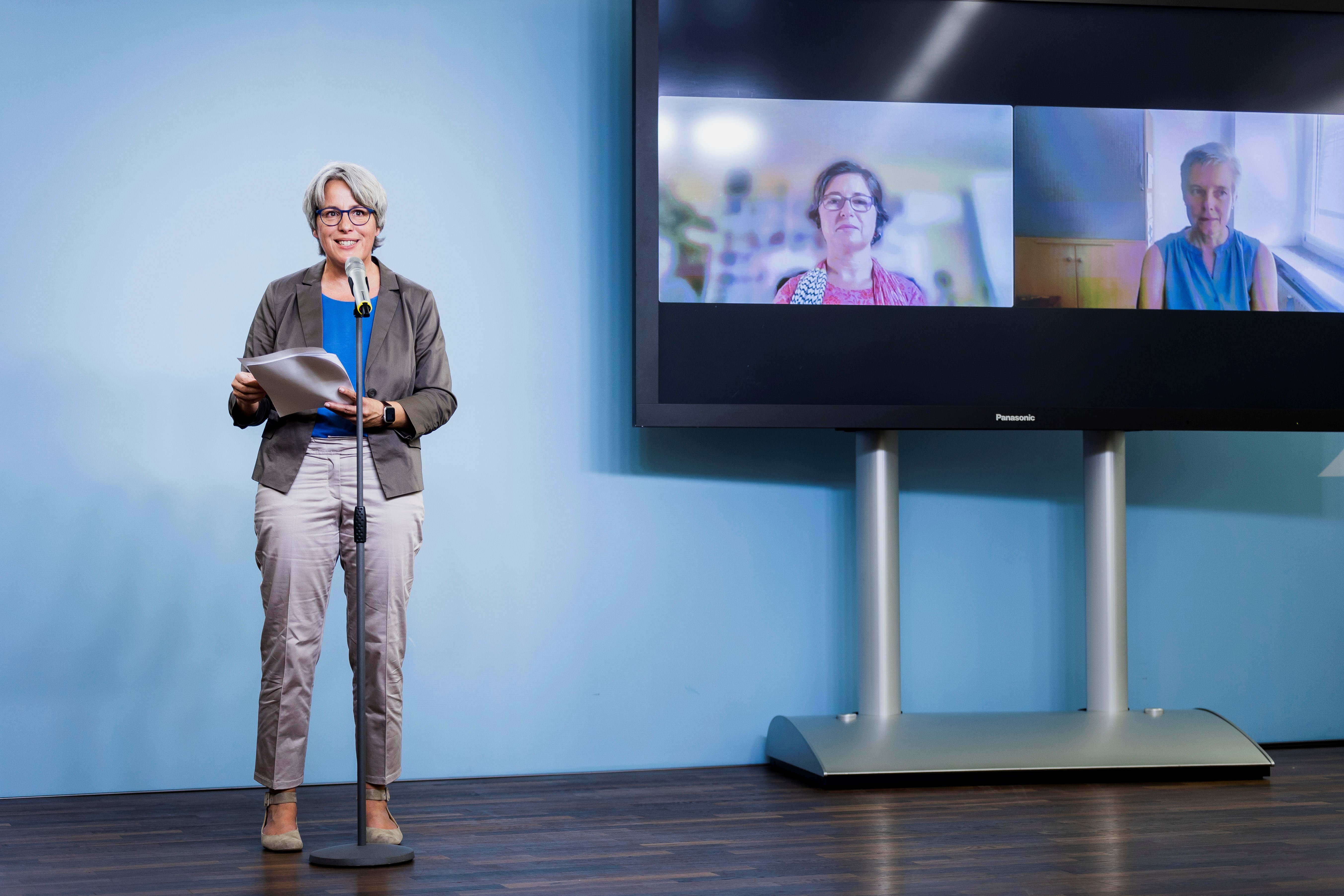 Kerstin Claus auf einer Bühne neben einem Monitor, hält eine Video-Konferenz ab.