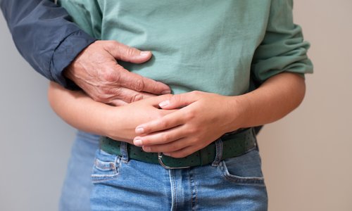 Ein Kind verschränkt die Arme vor dem Bauch, von hinten legt sich eine erwachsene Hand auf seinen Bauch.