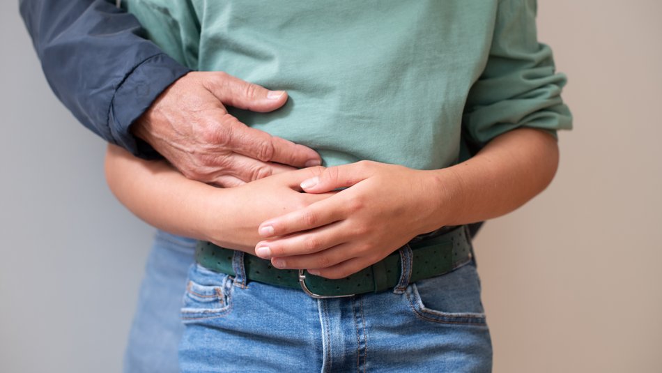 Ein Kind verschränkt die Arme vor dem Bauch, von hinten legt sich eine erwachsene Hand auf seinen Bauch.