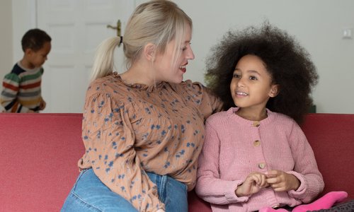 Eine Mutter führt auf dem Sofa ein Gespräch mit ihrer Tochter, im Hintergrund sieht man den kleinen Bruder