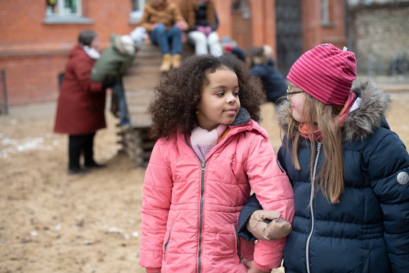 Zwei Kinder auf dem Spielplatz, im Hintergrund sieht man weitere Kinder und eine erwachsene Person