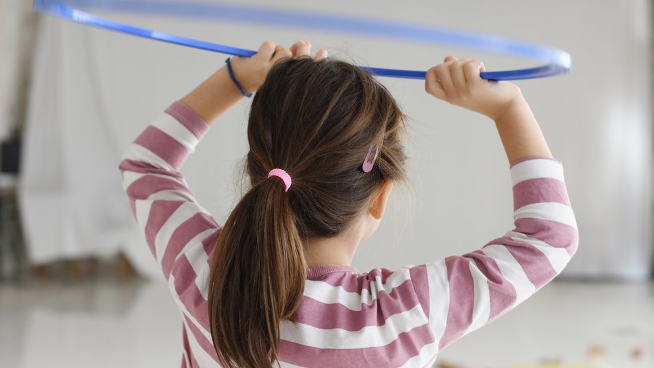 Das Foto zeigt ein Mädchen, das mit einem Hula-Hoop-Reifen spielt. Die Szene ist von hinten aufgenommen, das Gesicht des Mädchens ist von uns abgewandt.