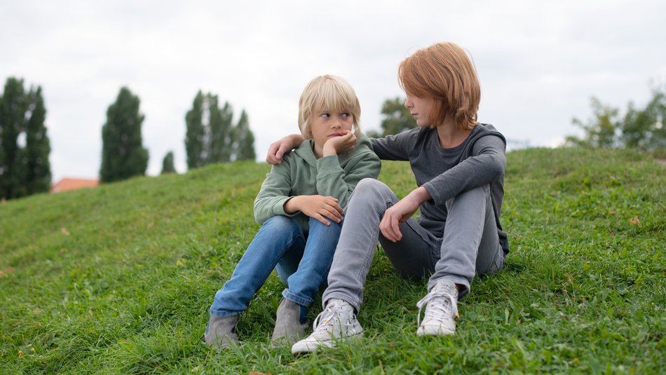 Zwei Jungen sitzen auf einer Wiese, der Ältere legt den Arm um den Jüngeren