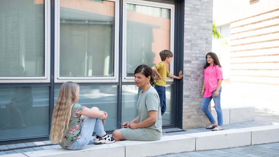 Vor einem Gebäude stehen zwei Kinder, ein weiteres Kind sitzt auf einem Mauervorsprung und unterhält sich mit einer erwachsenen Person.